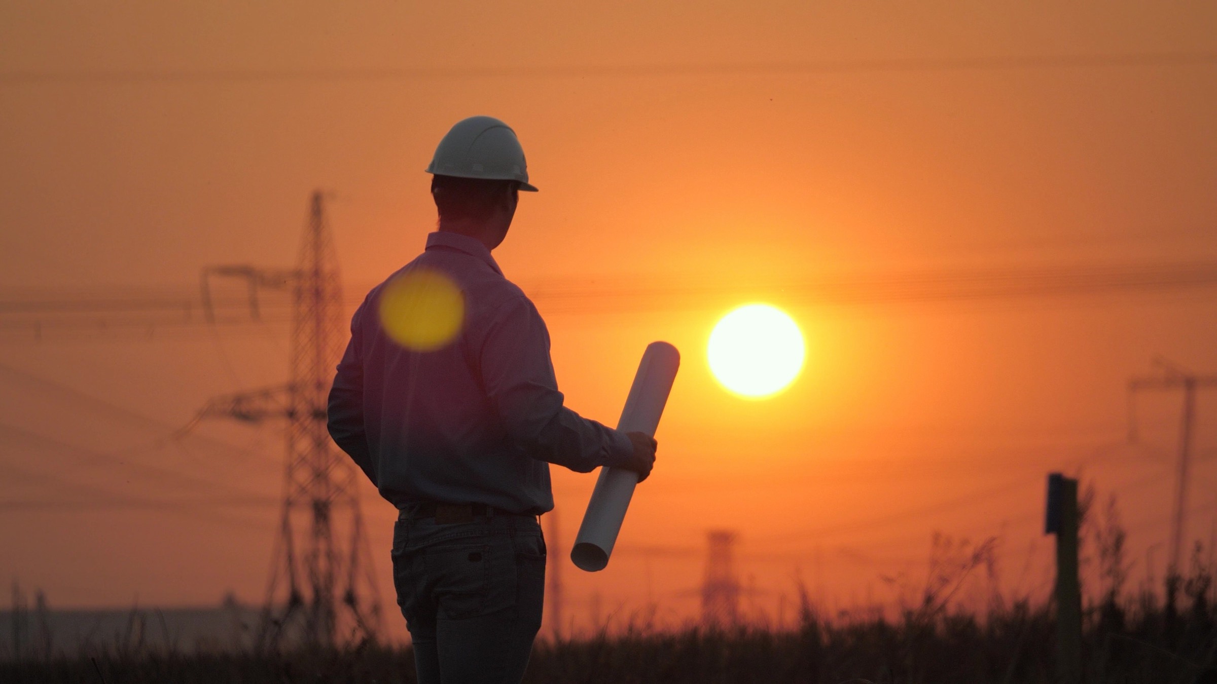 An engineer with rolled plans at dusk, transmission towers behind